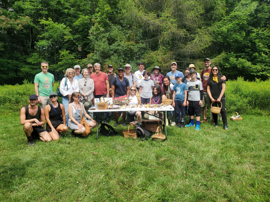 25 people of all different demographics smiling outside around a table full of mushrooms after a guided fungi forage through Catskill Fungi lead by John Michelotti.