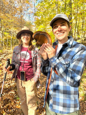 Mushroom walk participant smiling while holding a shelf mushroom in the Catskill Mountains. Yael Bernhard, walk leader of Catskill Fungi, looks at the mushroom and is also smiling as she  holds a walking stick and wears a Catskill Fungi Reishi T-Shirt.