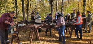 Sixteen people inoculating mushroom logs in Catskill fungi's outdoor mushroom cultivation space. Some people are drilling, plugging, waxing, and labeling logs to get them ready for incubation outside before they fruit shiitake mushrooms. 